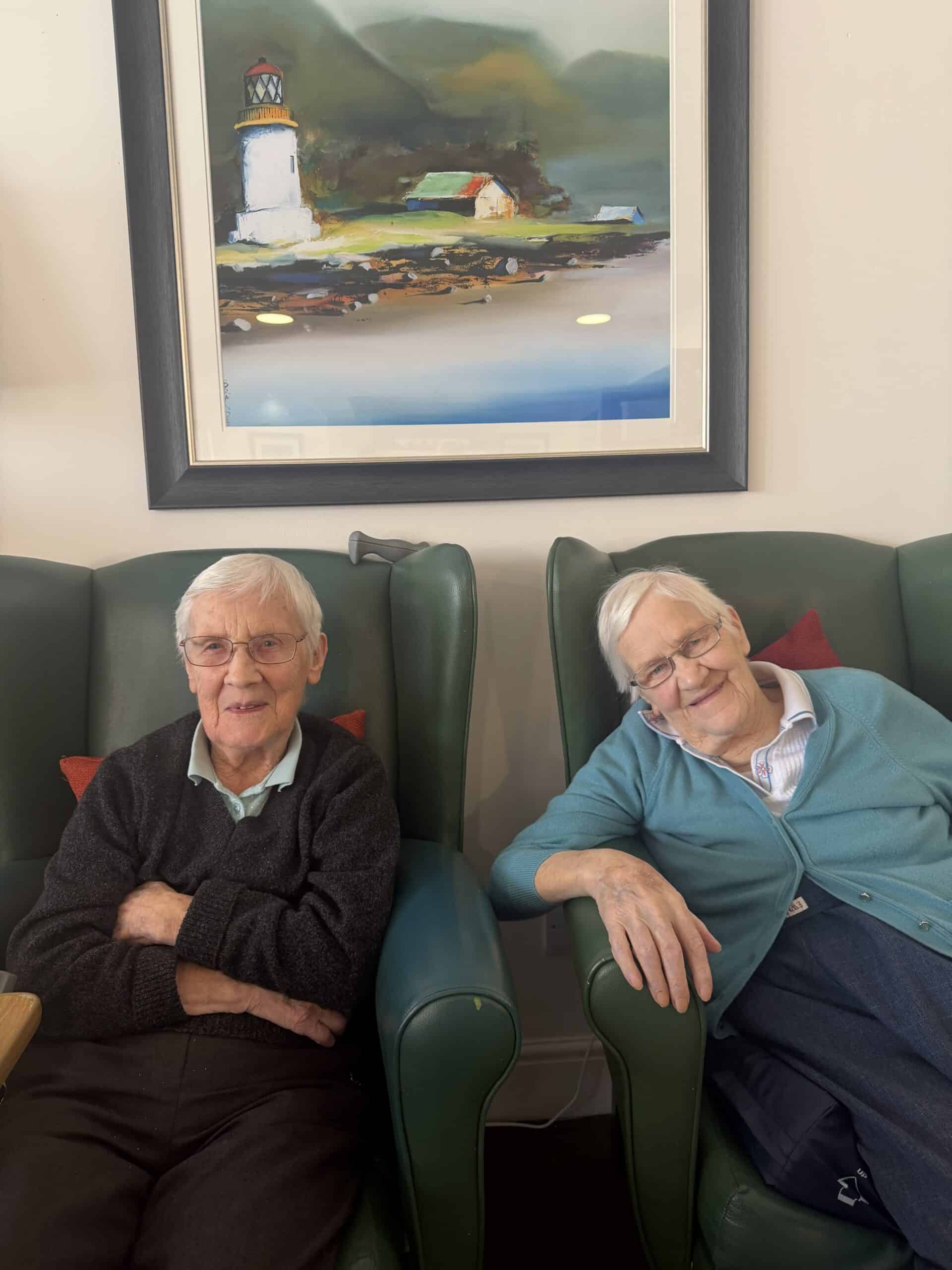 Two elderly residents smiling in a care home lounge with Dennis the Menace artwork behind them.