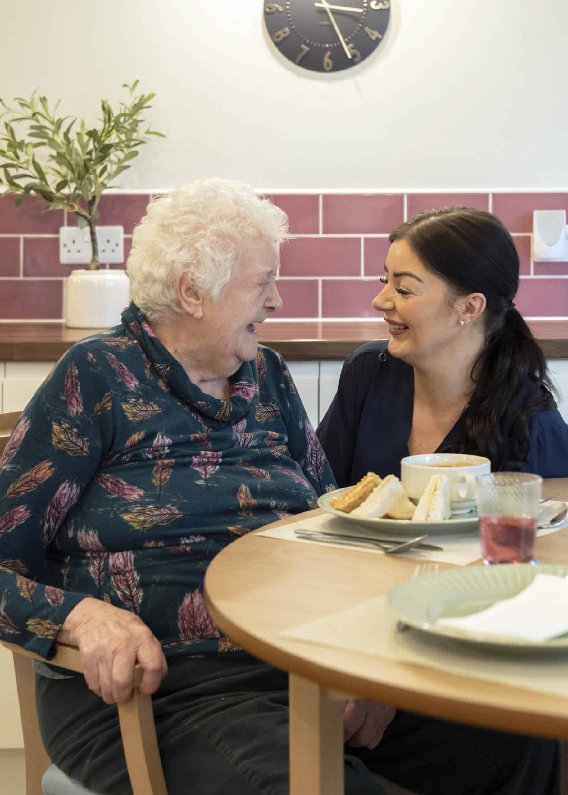 Elderly woman and caregiver sharing a joyful moment at the dining table.