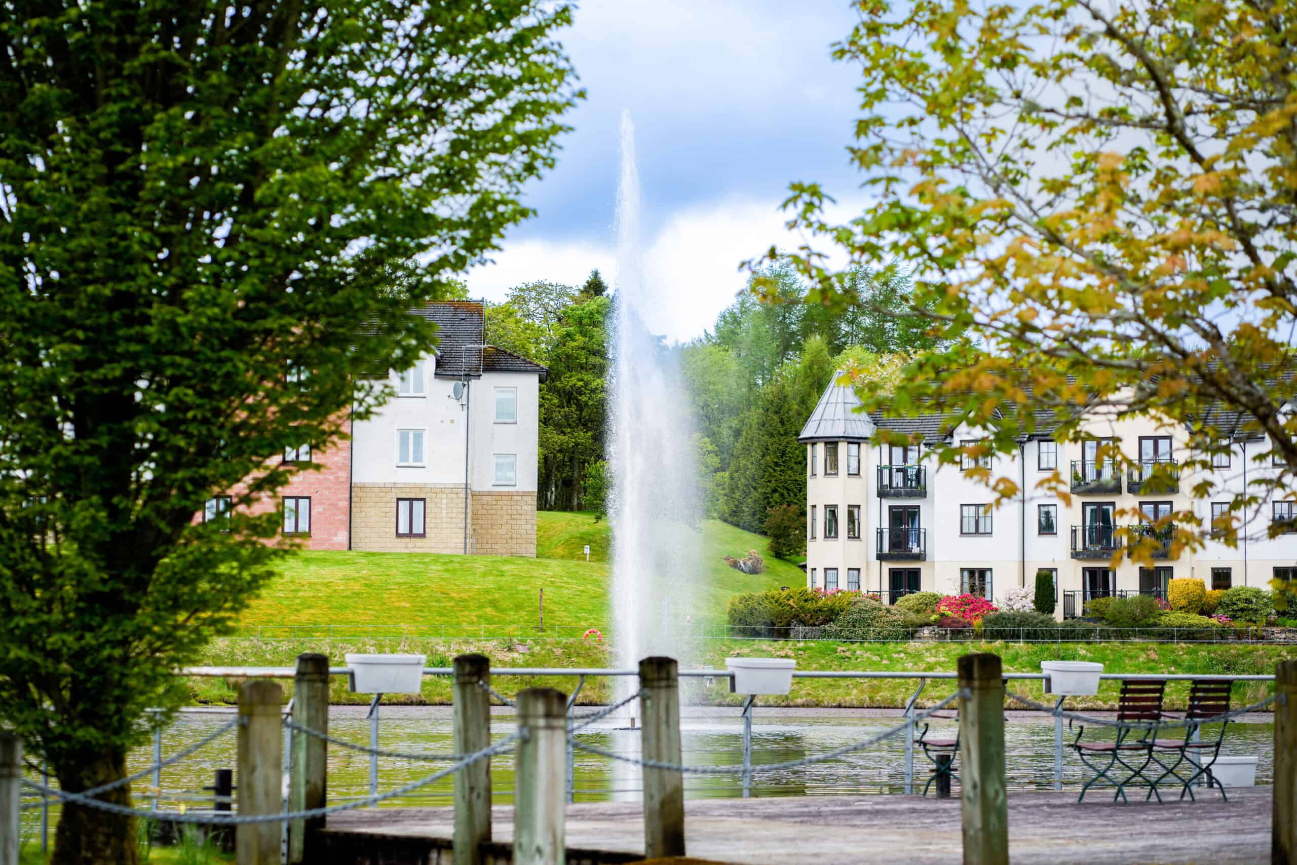 Park scene with fountain, trees, benches, and residential buildings in the background.